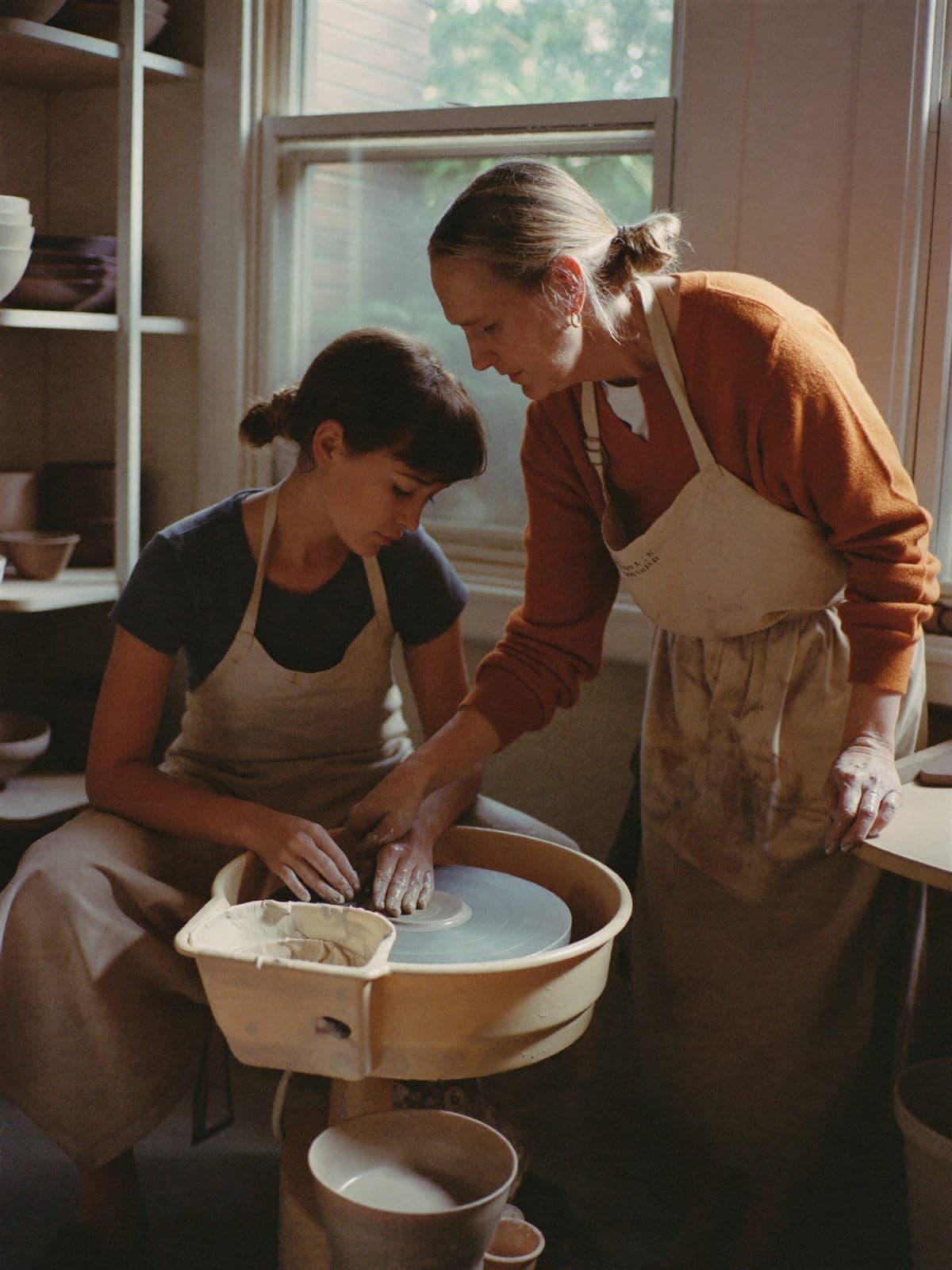 A ceramics teacher guiding a student's hands at the pottery wheel